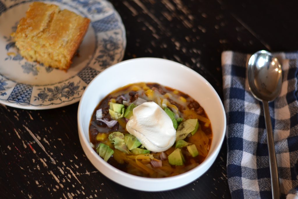 bowl of moose meat chili with avocado and dollop of sour cream on top with a napkin and spoon next to the bowl and slice of cornbread in the background