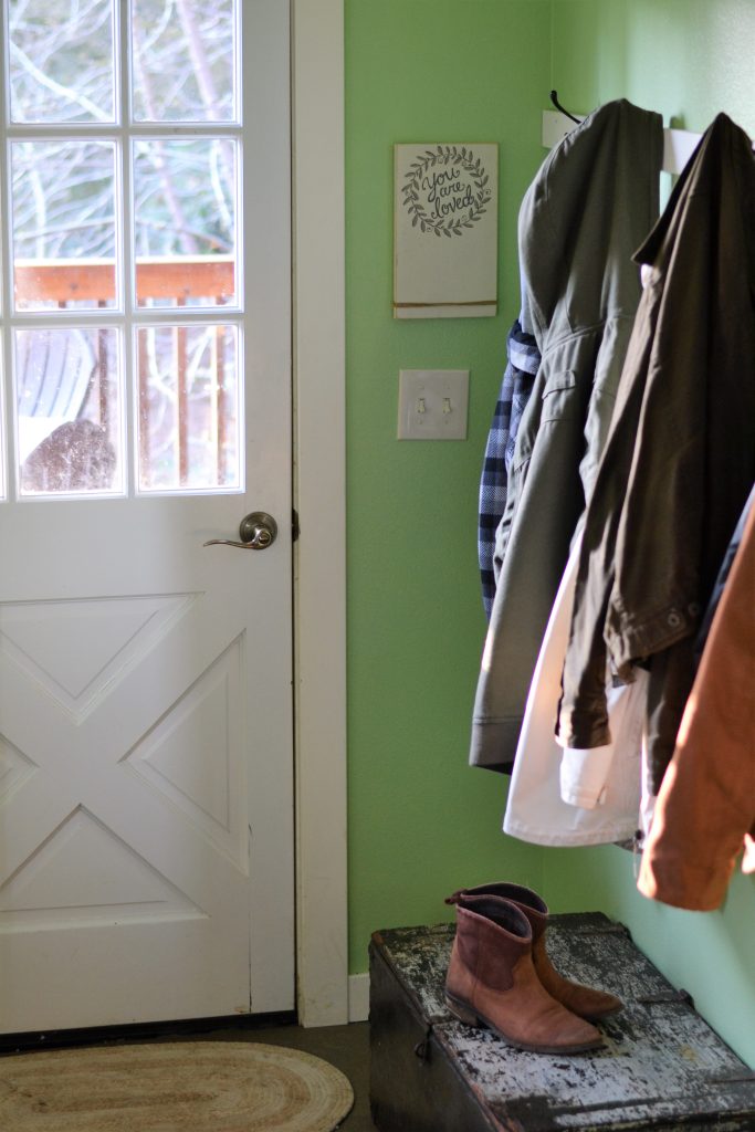 a room with a white door and bright green paint on the wall; a sign next to the door reads "you are loved"; coats hang next to the door and a pair of boots sits atop a large wooden box