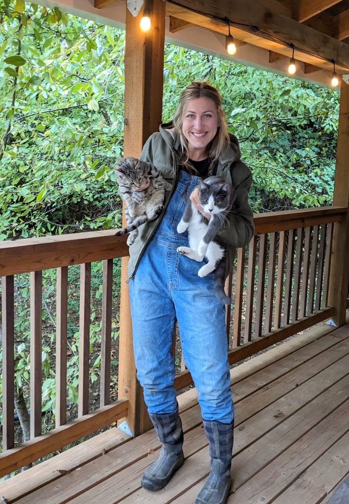 smiling girl in winter chore clothes- overalls and a work jacket and muck boots, holding two cats 